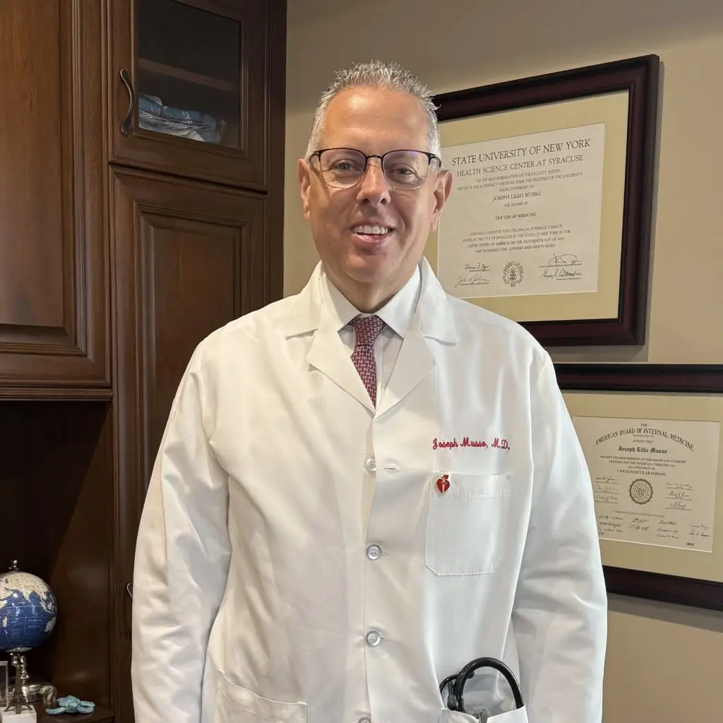 Comprehensive Cardiovascular PC - About: A man wearing a white lab coat stands in an office with framed diplomas on the wall and a wooden cabinet in the background, offering a glimpse into his professional achievements and expertise.